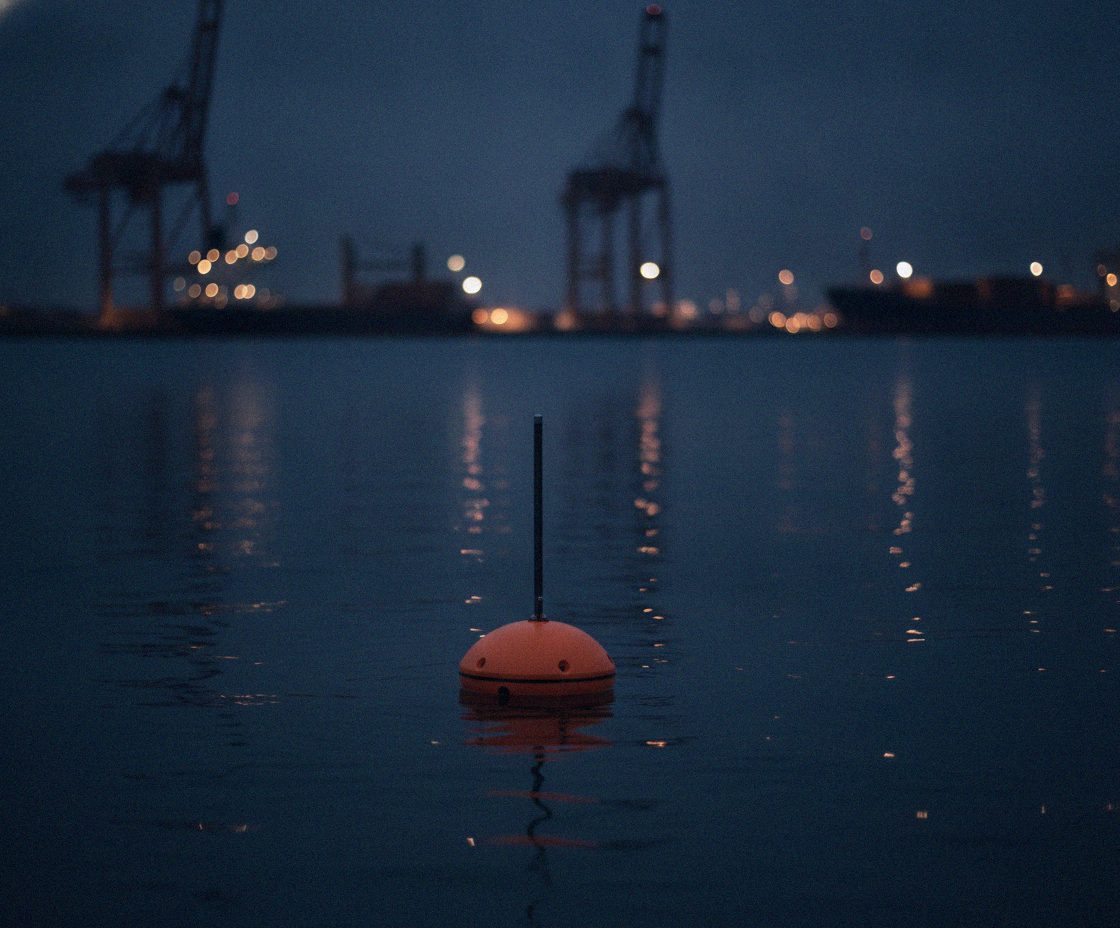 An Epsilon One sensor buoy in dark harbour water, port cranes in the background